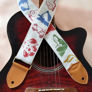 Puede incluir: Guitarra ac&uacute;stica con una correa blanca para guitarra con ilustraciones de perros coloridos. La correa tiene extremos de cuero color canela y herrajes color lat&oacute;n. La guitarra tiene un acabado rojo y negro.