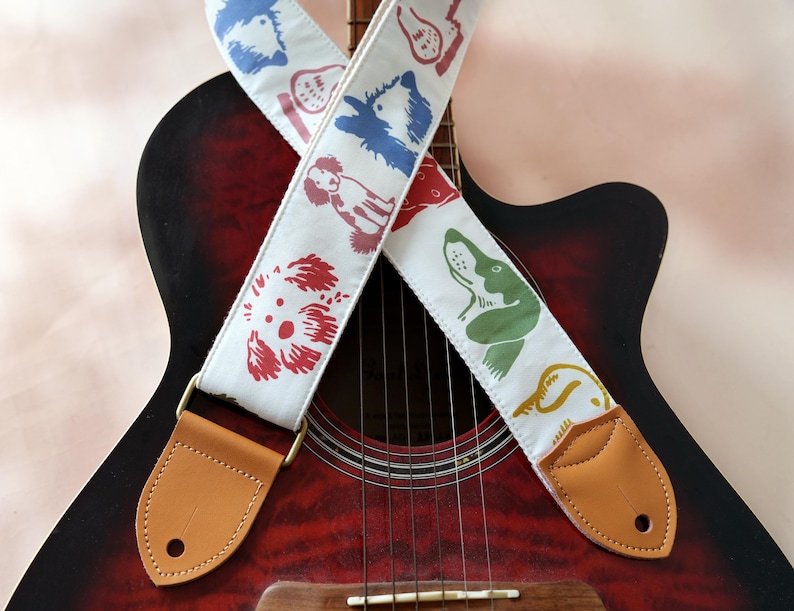 Puede incluir: Guitarra ac&uacute;stica con una correa blanca para guitarra con ilustraciones de perros coloridos. La correa tiene extremos de cuero color canela y herrajes color lat&oacute;n. La guitarra tiene un acabado rojo y negro.