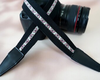 May include: Black camera strap with a white and pink butterfly pattern. The strap has black leather ends and is draped over a black camera with a lens.