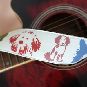 Puede incluir: Una correa de guitarra blanca con ilustraciones de perros coloridos en rojo, azul y verde. La correa se coloca contra una guitarra ac&uacute;stica roja, mostrando el dise&ntilde;o de la correa y los detalles del instrumento.