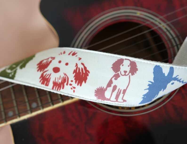 Puede incluir: Una correa de guitarra blanca con ilustraciones de perros coloridos en rojo, azul y verde. La correa se coloca contra una guitarra ac&uacute;stica roja, mostrando el dise&ntilde;o de la correa y los detalles del instrumento.