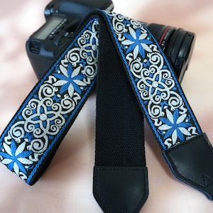 May include: A black camera strap featuring a blue and white floral embroidered pattern. The strap has a black canvas centre and black leather ends. The floral design includes blue star-shaped flowers and white scrollwork.