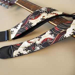 Pode incluir: Uma correia de guitarra com um padrão repetitivo de ondas oceânicas estilizadas em preto, branco e vermelho. A correia tem extremidades de couro preto com orifícios circulares para fixação e inclui uma fivela de ajuste de metal.