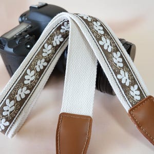 May include: A camera strap with a white canvas center and a decorative brown and white floral pattern. The strap has brown leather ends and is draped over a black camera.