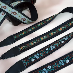 Peut inclure: Sangles d'appareil photo noires avec broderies florales colorées. Les sangles ont des extrémités en cuir noir et sont conçues pour être fixées à un appareil photo. Les motifs floraux incluent des fleurs orange, bleues et blanches.