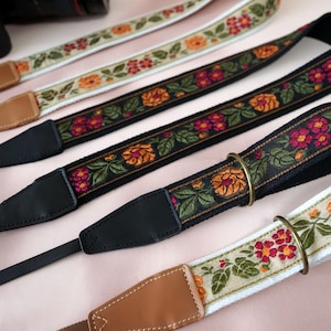 Puede incluir: Se muestran varias correas de cámara con bordados florales. Las correas presentan una variedad de colores, incluyendo negro, crema y blanco, con patrones florales naranjas, rojos y verdes. Las correas tienen cuero y herrajes metálicos.