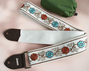 Könnte beinhalten: Weißer Gitarrengurt mit gestickten Blumen in Orange-, Blau- und Brauntönen. Der Gurt hat ein braunes Lederende mit dem aufgedruckten Wort "James".