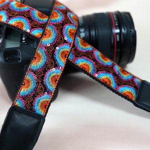 Pode incluir: Uma alça de câmera preta com um padrão bordado colorido de formas em leque laranja, azul e rosa. A alça é decorada com lantejoulas e tem extremidades de couro preto. A alça está sobre o corpo e a lente de uma câmera preta.