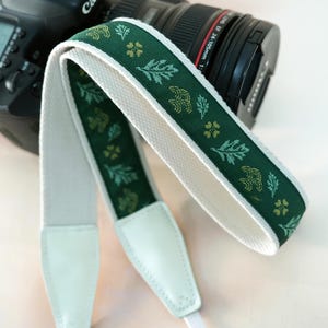 May include: A green and white camera strap with a floral pattern. The strap is made of a durable material and features white leather ends. The strap is designed to attach to a camera and is shown with a black camera body.