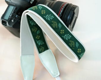 May include: A green and white camera strap with a floral pattern. The strap is made of a durable material and features white leather ends. The strap is designed to attach to a camera and is shown with a black camera body.