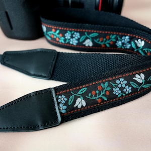 May include: Black camera strap with a floral embroidered design. The strap features a repeating pattern of white flowers, blue flowers, and red berries on a black background. The strap has black leather ends.