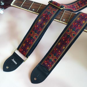 May include: A black guitar strap with a red, gold, and black paisley pattern. The strap has a leather end with the word "James" printed on it.
