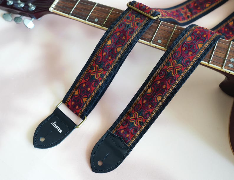 May include: A black guitar strap with a red, gold, and black paisley pattern. The strap has a leather end with the word "James" printed on it.