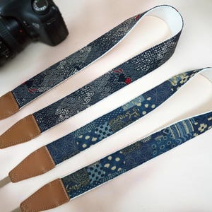 May include: Two patterned camera straps with brown leather ends. The straps feature a navy blue and white design with Japanese-inspired patterns. The straps are lying on a light pink surface, with a camera visible in the background.