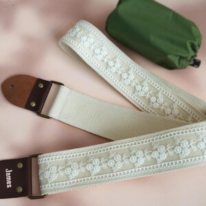 May include: A white cotton guitar strap with white embroidered floral details and brown leather ends. The strap has the word "James" printed on one end.