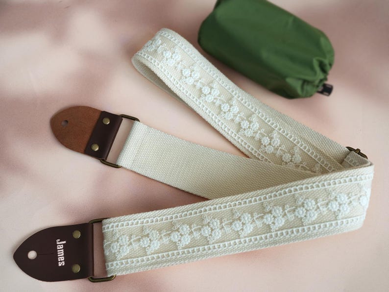 May include: A white cotton guitar strap with white embroidered floral details and brown leather ends. The strap has the word "James" printed on one end.