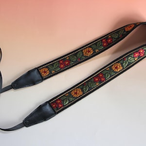 May include: A black camera strap with a floral design. The strap features a repeating pattern of red, yellow, and green flowers and leaves on a black background. The ends of the strap are black leather.