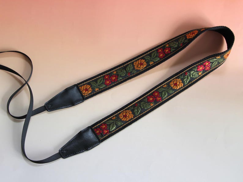 May include: A black camera strap with a floral design. The strap features a repeating pattern of red, yellow, and green flowers and leaves on a black background. The ends of the strap are black leather.