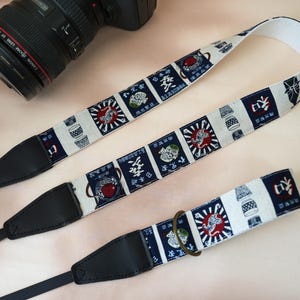 May include: A camera strap with a Japanese-inspired design. The strap features a repeating pattern of red, white, and blue squares with illustrations and Japanese characters. The strap has black leather ends and a black nylon strap.