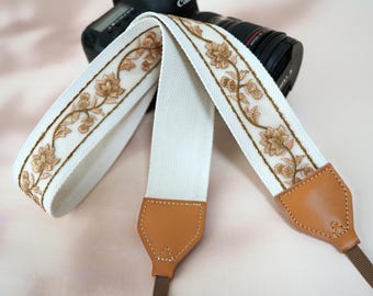 May include: Cream-colored camera strap with embroidered floral design in shades of brown. The strap features light brown leather ends and a brown strap connector. A black camera is visible in the background.