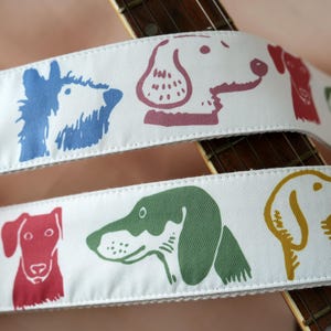 Puede incluir: Correa de guitarra blanca con ilustraciones de perros coloridos. La correa presenta un patr&oacute;n repetido de caras de perros en tonos de azul, rojo, verde y amarillo. La correa est&aacute; colocada sobre el m&aacute;stil de una guitarra.