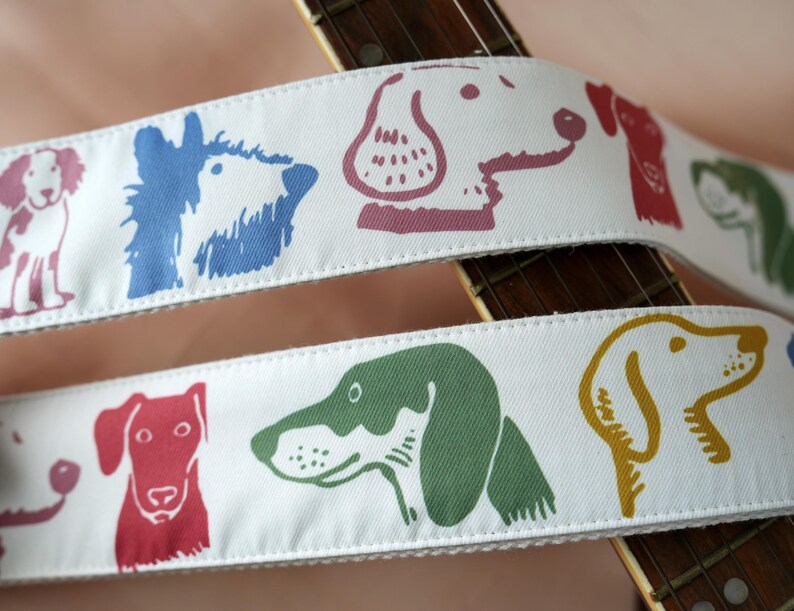 Puede incluir: Correa de guitarra blanca con ilustraciones de perros coloridos. La correa presenta un patr&oacute;n repetido de caras de perros en tonos de azul, rojo, verde y amarillo. La correa est&aacute; colocada sobre el m&aacute;stil de una guitarra.