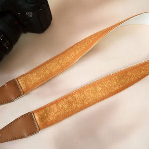 May include: A tan camera strap with a floral pattern in gold. The strap has brown leather ends and a white interior. A Canon EOS 7D camera is partially visible in the background.