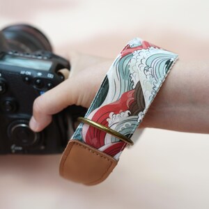 Può includere: Una tracolla per fotocamera con un motivo ripetuto di onde stilizzate rosse, bianche e verde acqua. La cinghia include un anello in metallo color ottone e terminali in similpelle marrone, fissata al corpo di una fotocamera digitale nera.