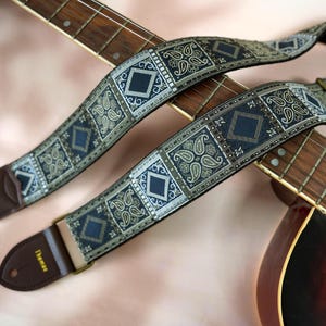 May include: Two guitar straps with a paisley and geometric pattern in navy blue, silver, and tan. The straps are positioned over the fretboard of a guitar. The straps have brown leather ends and metal hardware.