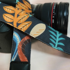 May include: A patterned camera strap with a dark blue background and a repeating design of orange leaves, blue and orange curved lines, and teal fern-like shapes. The strap is attached to a black camera body and lens.