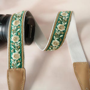May include: A white camera strap with a floral embroidered design in green, gold, and cream. The strap has tan leather ends and is attached to a black camera lens.