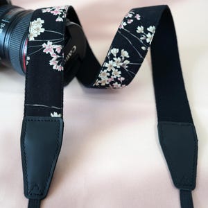 Pode incluir: Uma alça de câmera preta com um padrão floral em branco e rosa. A alça está presa a uma lente de câmera preta. A alça tem extremidades de couro preto e é projetada para conforto e estilo.