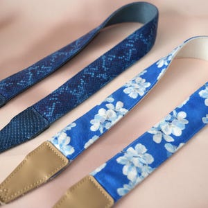 May include: Two camera straps. One is a dark blue snakeskin pattern, and the other is a vibrant blue with white floral print. Both straps have tan leather ends and are designed for cameras.