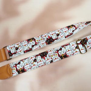 Peut inclure: Une sangle de guitare ornée d'un motif répété de chats porte-bonheur blancs, noirs et rouges. Elle est dotée d'extrémités en cuir marron et d'une boucle réglable de couleur laiton. Le fond est d'une couleur neutre et douce.