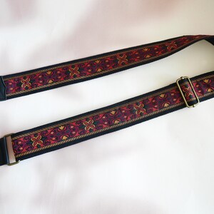May include: A black guitar strap with a red, gold, and black patterned design. The strap has a gold metal buckle and the word "James" is printed on the end.