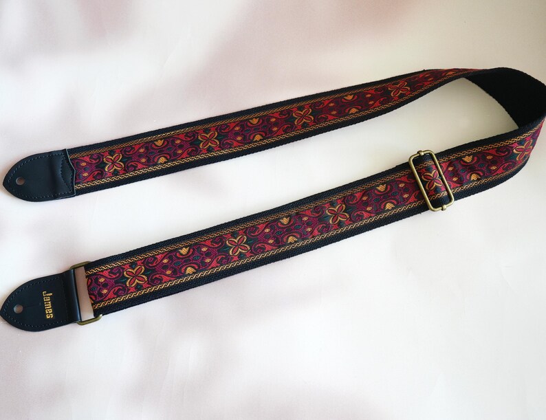 May include: A black guitar strap with a red, gold, and black patterned design. The strap has a gold metal buckle and the word "James" is printed on the end.
