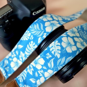 May include: A blue and white floral camera strap is wrapped around a black camera lens. The strap features a repeating pattern of white flowers and leaves on a blue background. A black Canon camera is visible in the background.