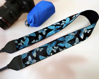 May include: A black camera strap with a repeating pattern of blue and silver butterflies. The strap is made of a woven material and has black leather ends. A blue camera bag and a camera are in the background.