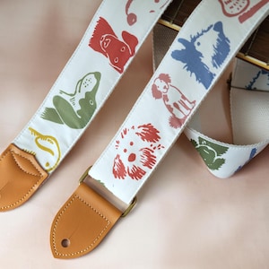 Puede incluir: Dos correas blancas para guitarra con ilustraciones coloridas de perros. Las correas tienen extremos de cuero marr&oacute;n y herrajes color lat&oacute;n. Las ilustraciones de perros son en rojo, verde, azul y amarillo. El m&aacute;stil de la guitarra es visible en el fondo.