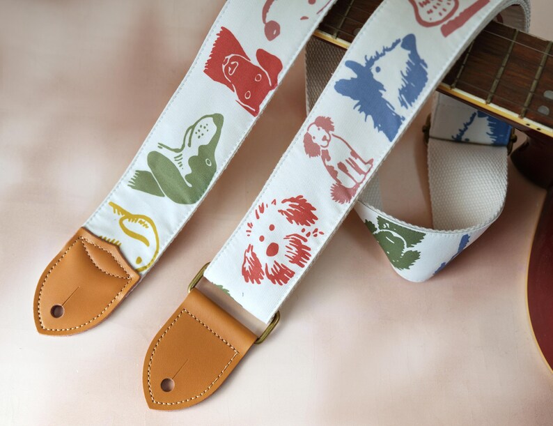 Puede incluir: Dos correas blancas para guitarra con ilustraciones coloridas de perros. Las correas tienen extremos de cuero marr&oacute;n y herrajes color lat&oacute;n. Las ilustraciones de perros son en rojo, verde, azul y amarillo. El m&aacute;stil de la guitarra es visible en el fondo.