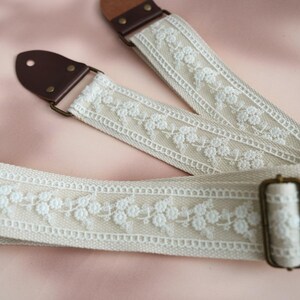 May include: A white guitar strap with a delicate floral lace design and brown leather ends. The strap has a brass buckle.