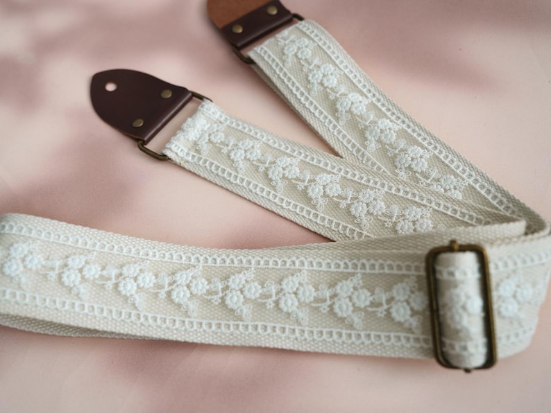 May include: A white guitar strap with a delicate floral lace design and brown leather ends. The strap has a brass buckle.