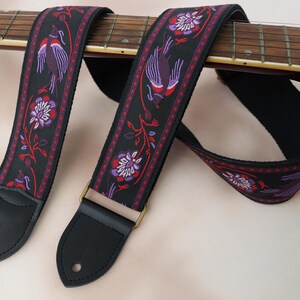 Puede incluir: Una correa de guitarra negra con un patrón repetitivo de pájaros y motivos florales en morado y rojo. La correa tiene extremos de cuero negro con orificios circulares para la fijación y una hebilla de metal color latón para ajustar la longitud.