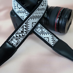 May include: Black and white camera strap with a geometric pattern. The strap is made of fabric and has a leather pad.