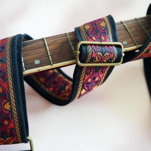 May include: A black guitar strap with a colorful patterned woven band. The strap has a brass buckle and is designed for use with a guitar.