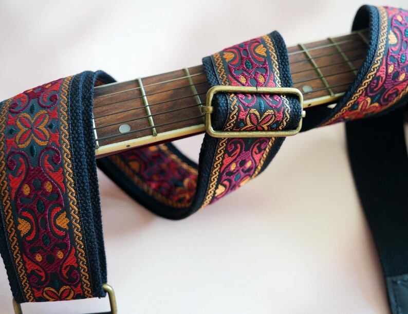 May include: A black guitar strap with a colorful patterned woven band. The strap has a brass buckle and is designed for use with a guitar.