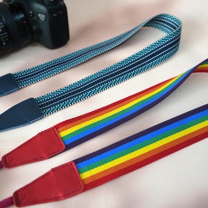 Op de afbeelding: Drie camerariemen worden getoond. Eén heeft een regenboogpatroon, de andere zijn blauw met witte en turquoise accenten. Alle riemen hebben leren uiteinden en zijn ontworpen om aan een camera te bevestigen.