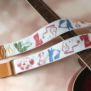 Puede incluir: Correa de guitarra blanca con ilustraciones coloridas de perros. La correa tiene extremos de cuero marr&oacute;n y herrajes met&aacute;licos dorados. Los dise&ntilde;os de perros son en rojo, azul, verde y otros colores. La correa se muestra en una guitarra ac&uacute;stica marr&oacute;n.