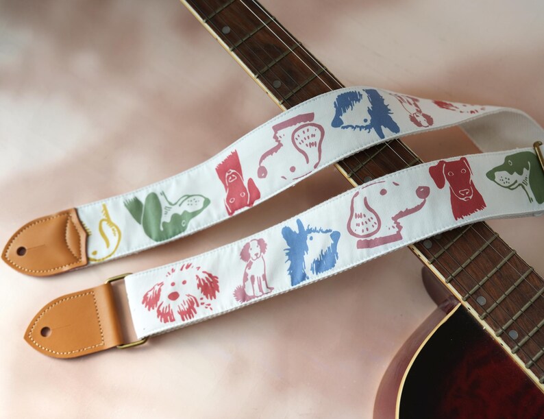 Puede incluir: Correa de guitarra blanca con ilustraciones coloridas de perros. La correa tiene extremos de cuero marr&oacute;n y herrajes met&aacute;licos dorados. Los dise&ntilde;os de perros son en rojo, azul, verde y otros colores. La correa se muestra en una guitarra ac&uacute;stica marr&oacute;n.
