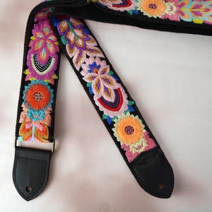 Pode incluir: Correia de guitarra preta com bordado floral vibrante em tons de laranja, rosa, roxo, azul e verde. A correia apresenta extremidades em couro e ferragens metálicas. Um acessório decorativo para instrumentos musicais.